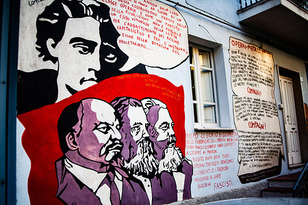 A mural painting of Marxist philosopher Antonio Gramsci (top left) with the Socialist thinkers (L-R) Vladimir Lenin, Karl Marx, and Friedrich Engels on a wall in downtown. The Sardinian town of Orgosolo is renowned as an open-air museum, with over 150 murals adorning its historic center that reflect local culture, pastoral heritage, and global political struggles. Influenced heavily by Cubism and Social Realism, the murals use a bold, graphic style to depict scenes of shepherd life, social justice movements, and anti-war protest.