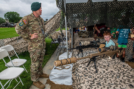 A U.S. Army member stands and watches as a child poses for a photo with a sniper rifle during a public event. Thousands of U.S. Army members have arrived in Washington D.C. for a display of their equipment and a parade around the Capitol Mall, commemorating its 250th anniversary.