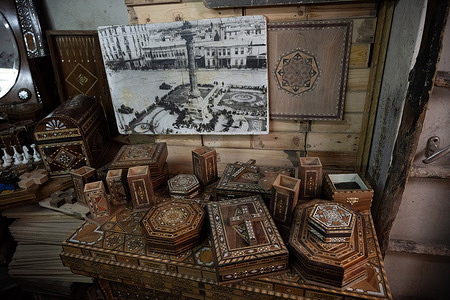 A display of inlaid wooden mosaic boxes , an old black and white photograph of Merjeh Square seen inside the Workshop of Syrian artisan Abu Toni. The art of Damascus mosaic has declined in recent years due to the emigration of artisans and the economic effects of conflict. Despite these challenges, it remains a symbol of Syrian heritage.