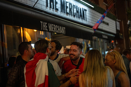 A fan wrapped in England flag seen interacting with other England fans beneath the awning of The Merchant during England's victory celebrations. England fans celebrate in Brentwood High Street following England's victory against Norway in the World Cup.