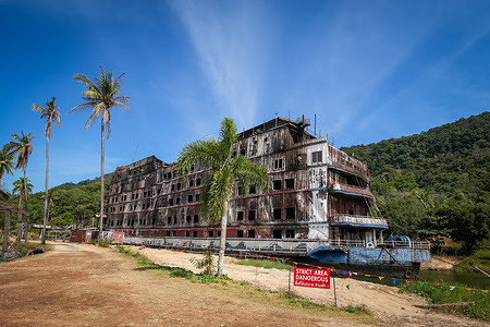 A red warning sign reading ‘Strict Area Dangerous’ seen at the Koh Chang Grand Lagoona Resort on Koh Chang Island. The abandoned Ghost Ship hotel of Koh Chang Island known as the Galaxy and owned by Thai land magnate Oran Asawaritthikul was a seven-storey luxury ship hotel with around 200 rooms. Opened around 2004 as the centerpiece of the upscale Koh Chang Grand Lagoona Resort. The resort closed around 2016 due to economic challenges and legal issues while the ship’s eerie, haunting allure turned it into a popular urban-exploration site. In December 2024, a fire fueled by winds and the wooden structure destroyed much of the ship. The exact cause of the fire remains undetermined. With the remaining structure deemed unsafe, the upper floors were dismantled by mid-2025 and sold for scrap.