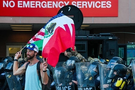 A protestor flashes a peace sign while holding a hybrid U.S.–Mexico flag in front of a line of riot police and tactical officers during an anti-ICE demonstration. Demonstrators staged a protest as part of a citywide action against U.S. Immigration and Customs Enforcement (ICE), where protesters called for the end of deportations, the shutdown of detention centers, and protection for migrant communities across the United States.