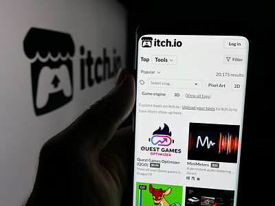 In this photo illustration, a person is holding a smartphone with the web page of indie video game company itch.io on screen in front of a logo.