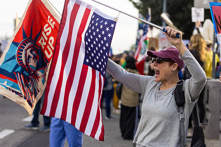 A protester waves a U.S. flag and shouts during an anti-ICE demonstration outside the Metropolitan Detention Center in Los Angeles, part of a national escalation following immigration enforcement actions and unrest linked to ICE activity in Minnesota.