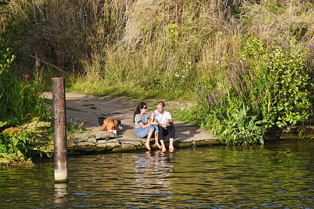 A couple sitting at the edge of the River Thames dipping their feet in the water while enjoying a drink at Hampton Court as the weather reaches 30°.