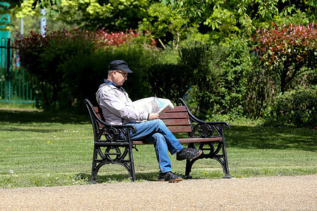 A man sitting on a park bench reads a newspaper on a warm day in London.