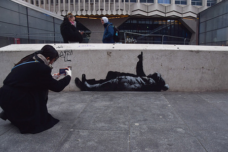 A woman takes a photo of the new Banksy artwork on a wall outside Centre Point and Tottenham Court Road station. Two identical new artworks, one of which has now been confirmed by elusive street artist Banksy, have appeared outside Centre Point and on an unoccupied mews building in Bayswater. The artworks depict two children lying on their backs and one child pointing upwards.
