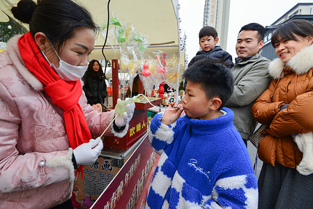 A boy blows a sugar figurine at a Spring Festival market stall, with a craftswoman assisting and onlookers watching.