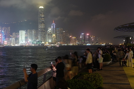 People gather along the waterfront of Tsim Sha Tsui to watch the night view of Hong Kong Island and Victoria Harbour in Hong Kong. Along the Tsim Sha Tsui waterfront, the evening transitions into a shared public ritual as the Hong Kong Island skyline begins to illuminate. Locals and travelers from around the world gather along the Avenue of Stars, finding their own pockets of space to witness the city's nightly transformation. Some lean against the railings to watch the Star Ferry cross the harbour, while others navigate tripods and smartphones to capture the shifting light. It is a moment defined by a collective, quiet observation, where the scale of the architecture meets the steady, everyday movement of the crowd along the water's edge.
