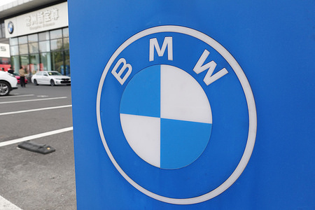 A close-up of the BMW logo on a blue sign, with a BMW dealership and cars in the background.