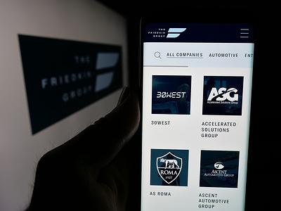 In this photo illustration, a person is holding a smartphone with the website of US company The Friedkin Group on screen in front of logo.