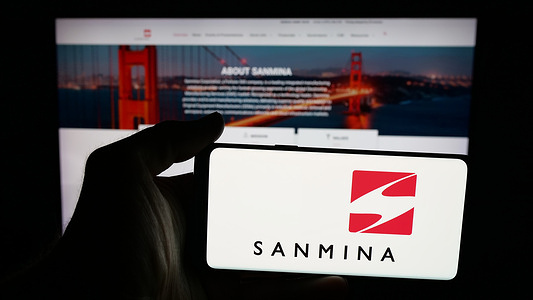In this photo illustration, a person is holding a mobile phone with the logo of US electronics company Sanmina Corporation on screen in front of web page.
