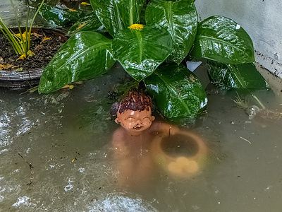 A smiling Thai clay child statue holding a jar, partially submerged in murky floodwater up to its mouth, in a garden in Lat Phrao District. All 50 districts of Bangkok, Thailand, were declared emergency disaster zones, following torrential monsoon downpours over a 48-hour period. Lat Phrao District is one of the hardest-hit areas in Bangkok.