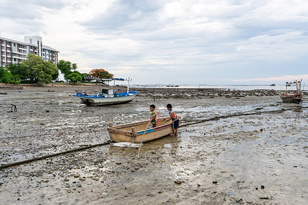 Young boys seen in a small wooden boat resting on a muddy shoreline with debris exposed during low tide in a fishing community in Bang Phra. Bang Phra is on the eastern coast in Chonburi province is home to a local fishing community. During low tide, exposed mudflats reveal waste trapped along the shoreline while fishing boats rest on the mud as fishermen continue their daily activities.