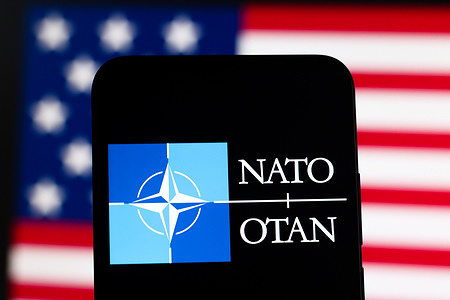 In this photo illustration, the North Atlantic Treaty Organization (NATO - OTAN) logo is seen displayed on a smartphone screen with the flag of the United States in the background.