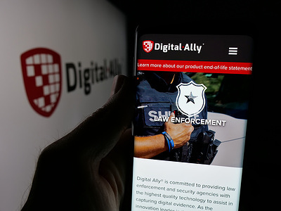 In this photo illustration, a person is holding a cellphone with the webpage of US video solutions company Digital Ally Inc. on screen in front of logo.