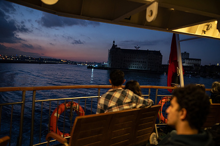 Passengers enjoy the view on a city line ferry at sunset in Istanbul, with Haydarpasa Train Station in the background.