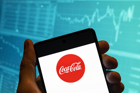 In this photo illustration, the American soft drink brand Coca-Cola logo is seen displayed on a smartphone with an economic stock exchange index graph in the background.