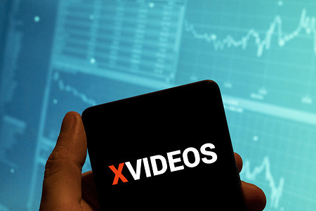 In this photo illustration, the adult pornographic video-sharing website Xvideos logo is seen displayed on a smartphone with an economic stock exchange index graph in the background.