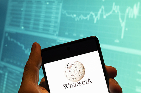 In this photo illustration, the multilingual, web-based, free encyclopedia Wikipedia logo seen displayed on a smartphone with an economic stock exchange index graph in the background.