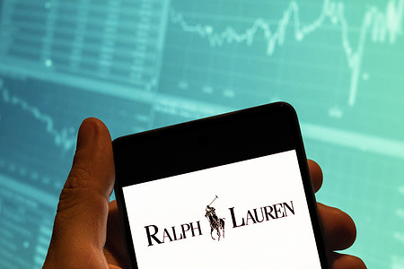 In this photo illustration, the American fashion brand Ralph Lauren logo is seen displayed on a smartphone with an economic stock exchange index graph in the background.