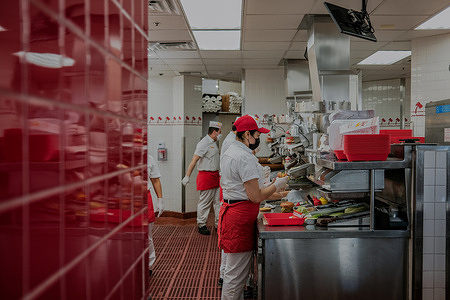 A behind-the-scenes shot of an In-N-Out kitchen, with employees grilling fresh patties and assembling burgers with lightning speed. TThe kitchen is clean and well-organized, with large signs reminding employees of the company's commitment to quality.