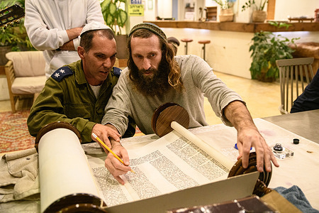 Member of the Kfar Azza community sits next to a volunteer writing a bible scroll in honor of the 7th October hostages and victims. Members of Kibbutz Kfar Azza gathered to watch the screening of the kibbutz women and kids release from Hamas captivity after they were taken hostages from their houses on October 7th.