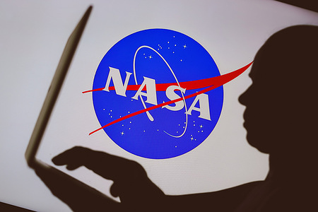In this photo illustration, the National Aeronautics and Space Administration (NASA) logo is seen in the background of a silhouette of a person using a notebook.