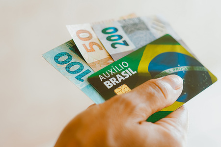 In this photo illustration, a hand seen holding the Auxílio Brasil ATM card (Bolsa Família) with 100, 200, and 50 Brazillian bank notes. The Brazilian Cash Transfer Program, known as Bolsa Família or Auxílio Brasil, is one of the largest conditional cash transfer programs globally. It focuses on providing financial aid to low-income and marginalized families in an effort to alleviate poverty and eradicate hunger. The program operates by offering monthly cash assistance to eligible households.