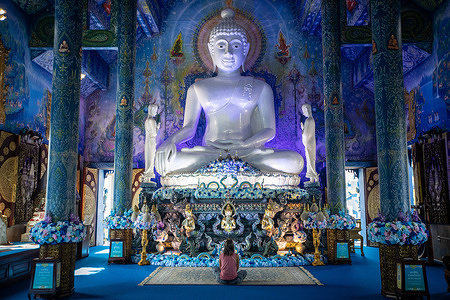 A Western tourist sits in front of the white sitting Buddha statue inside the Blue Temple. Wat Rong Suea Ten (Temple of the Dancing Tiger), most commonly known as the "Blue Temple" is a fusion of traditional Buddhist values and classic Thai architecture with contemporary design choices, it was designed by Putha Kabkaew, a student of the artist, Chalermchai Kositpipat, who built the very well known "White Temple".