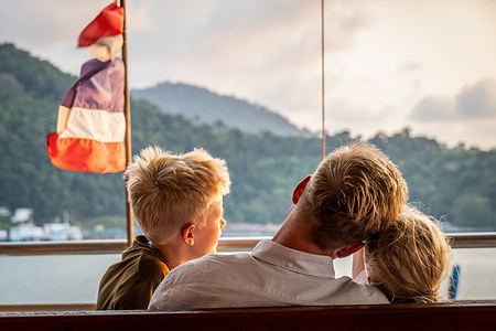 A Scandinavian tourist is seen with his children watching the sunset on a ferry on the way to Koh Chang Island, with a Thai flag in the background. Tourists exploring Thailand have a range of transportation options at their disposal, from buses and trains to subways, vans, ferries, and iconic Song Taews (small pick-up trucks with two rows of benches).