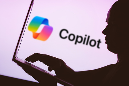 In this photo illustration, the Microsoft Copilot logo is seen in the background next to a silhouette of a person using a notebook.