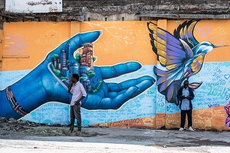 Two men stand in front of murals depicting a hand holding a city and a flying bird painted on a wall in Nakuru Town, created as part of Nakuru's UNESCO Creative City project to showcase the city's cultural and artistic heritage. This collaborative work features contributions from various graffiti artists including Bankslave, Joel Bergner, Chelwek, Dante and Natasha Floortje.