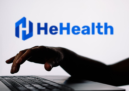 In this photo illustration, the HeHealth logo is seen in the background of a silhouette of a person using a notebook.