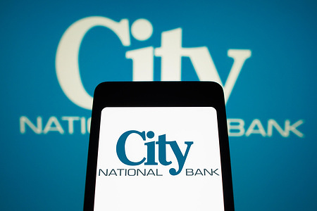In this photo illustration, the City National Bank logo is displayed on a smartphone screen and in the background.