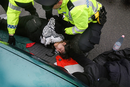 An activist from Palestine Action is arrested after being cut out of a lock in Discovery Park, while being watched over by police officers. Protesters staged a lie-in protest, blocking the three access roads to Discovery Park, a business estate. Instro Precision, a company owned by Israeli arms company Elbit Systems, operates a factory producing sights for Israeli sniper rifles. The demonstrators argue that these weapons are used against Palestinians in Gaza and other places. This action was part of the A15 coordinated global economic blockade for Palestine.