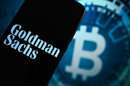 In this photo illustration, a Goldman Sachs logo is displayed on a smartphone with a Bitcoin logo in the background.