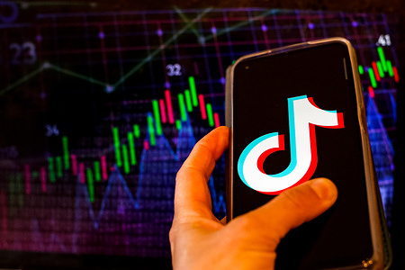 In this photo illustration, the TikTok logo is seen displayed on a smartphone screen against a screen displaying a trade markets illustration.