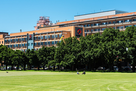 View of the Alfred hospital from the Fawkner park across the mowed lawn, with people at the park. 
Alfred hospital in Melbourne is one of the central hospitals with General hospital, ER and specialist medical suites as well as a major medical research precinct.