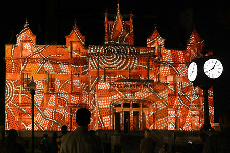 A man is silhouetted while watching a projection mapping art show.Jeff Dobrow brought the art of 12 Australian Aboriginal Artists and musicians for an art installation using Projection Mapping on the side of a building. The art show is run from a small laptop and was replayed two nights in a row. The show was called Light the Way Forward.