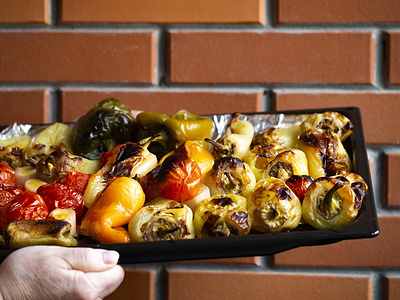 Baked vegetables consisting of bell peppers, tomatoes and leek on a tray with a brick wall background.