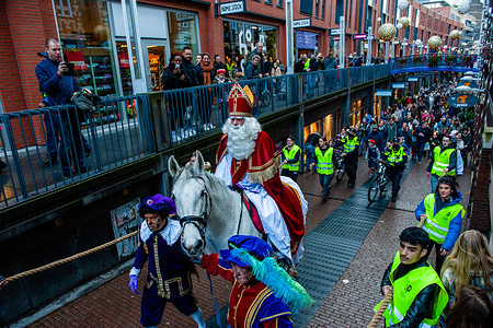 St. Nicholas is seen on the street riding his white horse called, Ozosnel.
The first Saturday after November 11th, the red-and-white-clad Sinterklaas (St. Nicholas) arrive with great fanfare to several Dutch cities. In Nijmegen, the white-bearded legend makes his entrance into the city riding his white horse through the city center. After years of protest against the traditional form of Zwarte Piet or Black Pete, a St Nicholas’ helper who usually appears as a blackface character with large gold earrings and exaggerated lips, last year the organization decided change this look, and introduced new helpers without blackface makeup, but still wearing Afro black wigs.