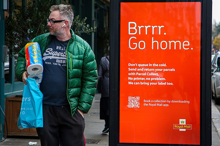 A man stands next to a digital advertisement of the Post Office in London.