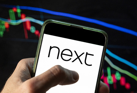 In this photo illustration the British multinational clothing, footwear and home products retailer Next plc logo seen displayed on a smartphone with an economic stock exchange index graph in the background.