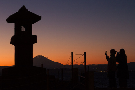 Silhouettes of people taking photos of Mount Fuji at sunset while standing next to a stone lantern on Enoshima Island.